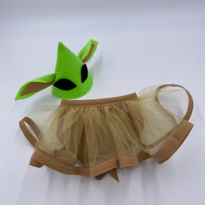 May include: A green alien-themed hat with black eye cutouts and brown ear details, paired with a tan tulle skirt. The skirt has a brown trim and a bow in the back. The costume set is designed for a child.