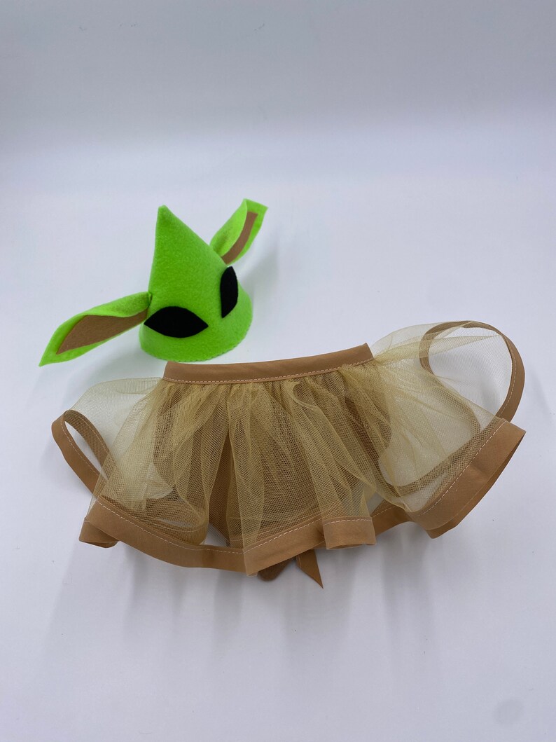 May include: A green alien-themed hat with black eye cutouts and brown ear details, paired with a tan tulle skirt. The skirt has a brown trim and a bow in the back. The costume set is designed for a child.