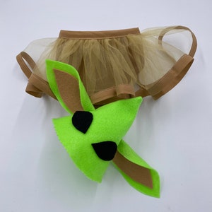 May include: A green felt character head with black eye accents and long ears, paired with a tan tulle skirt with a brown trim. The costume is designed for a pet.