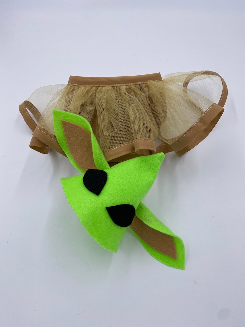 May include: A green felt character head with black eye accents and long ears, paired with a tan tulle skirt with a brown trim. The costume is designed for a pet.