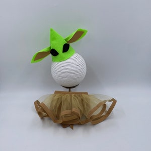 May include: A green felt hat with large ears and black eye shapes, paired with a tan tutu. The hat sits atop a white, rope-covered sphere. The tutu has a tan trim and a bow.
