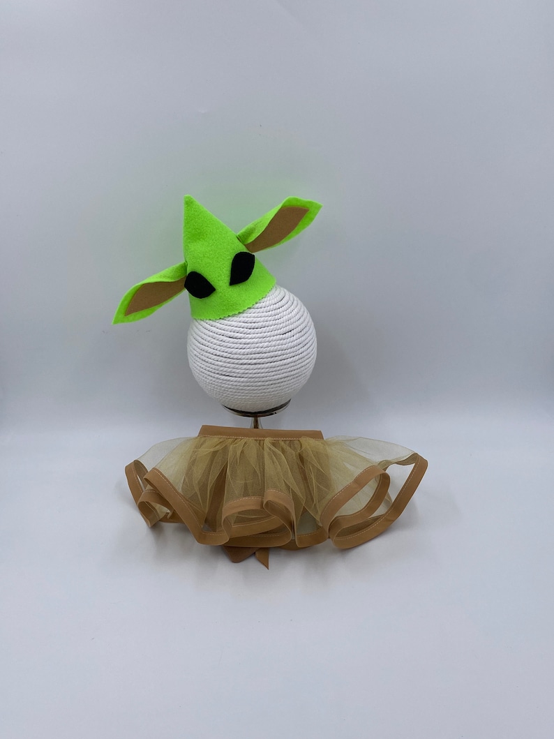 May include: A green felt hat with large ears and black eye shapes, paired with a tan tutu. The hat sits atop a white, rope-covered sphere. The tutu has a tan trim and a bow.