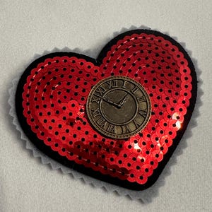 May include: Red heart-shaped sequin patch with a black border and a gold clock face in the center. The clock face has Roman numerals and black hands.