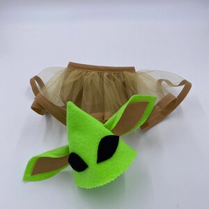 May include: A green felt alien costume head with large black eyes and brown felt ears, paired with a tan tulle skirt. The costume is designed for pets, likely a dog or cat, and is set against a white background.