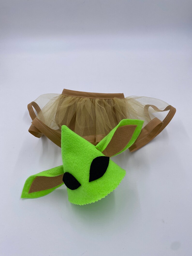 May include: A green felt alien costume head with large black eyes and brown felt ears, paired with a tan tulle skirt. The costume is designed for pets, likely a dog or cat, and is set against a white background.