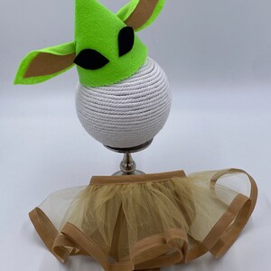 May include: A green felt hat with brown ears and black eyes sits atop a white rope-wrapped sphere. Below, a tan tulle skirt with a brown trim and bow completes the costume set.