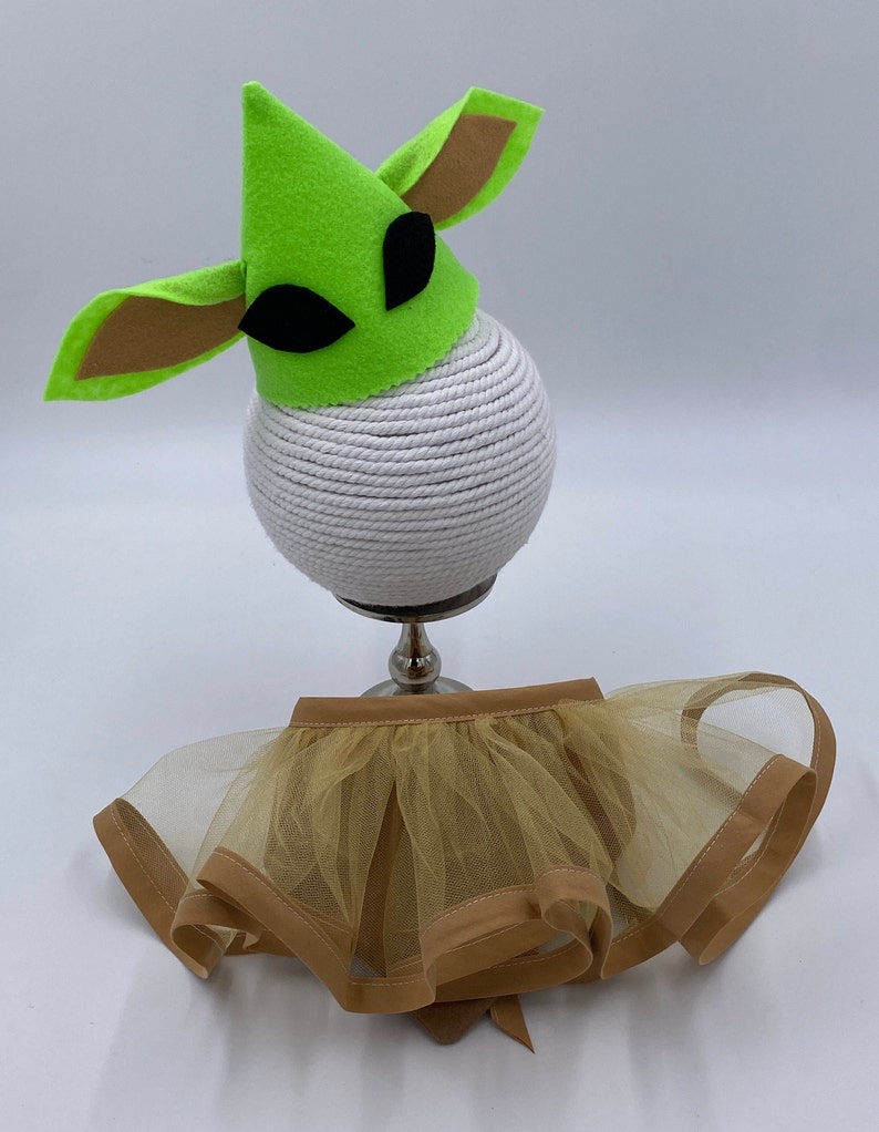 May include: A green felt hat with brown ears and black eyes sits atop a white rope-wrapped sphere. Below, a tan tulle skirt with a brown trim and bow completes the costume set.