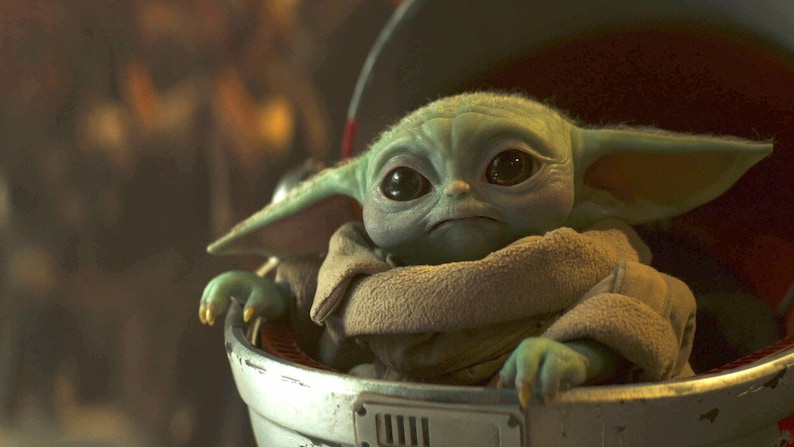 May include: A close-up of a green alien-like creature with large black eyes and pointed ears, wrapped in a tan blanket. The creature is in a silver and red carrier, with a serious expression. The image is from the Star Wars franchise.