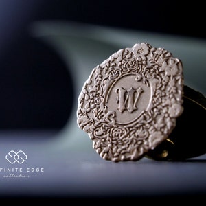 May include: A round, cream-colored, decorative seal with an intricate floral design and the letter "W" in the center. The seal is made of metal and has a slightly textured surface.