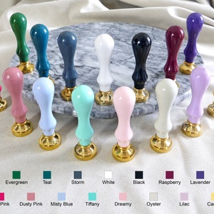 Pu&ograve; includere: Una collezione di 18 timbri per sigilli di cera colorati con basi dorate. I timbri sono disposti in cerchio su una superficie di marmo. I colori includono verde smeraldo, verde acqua, tempesta, bianco, nero, lampone, lavanda, rosa acceso, rosa tenue, blu nebbioso, tiffany, da sogno, ostriche, lilla e canarino.
