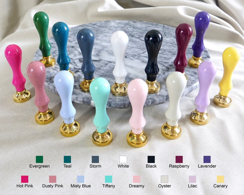 Pu&ograve; includere: Una collezione di 18 timbri per sigilli di cera colorati con basi dorate. I timbri sono disposti in cerchio su una superficie di marmo. I colori includono verde smeraldo, verde acqua, tempesta, bianco, nero, lampone, lavanda, rosa acceso, rosa tenue, blu nebbioso, tiffany, da sogno, ostriche, lilla e canarino.