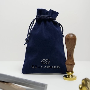 Pu&ograve; includere: Una pochette in velluto blu navy con chiusura a cordoncino e il testo "GETMARKED" in bianco. La pochette &egrave; accanto a un timbro in legno e oro con manico.