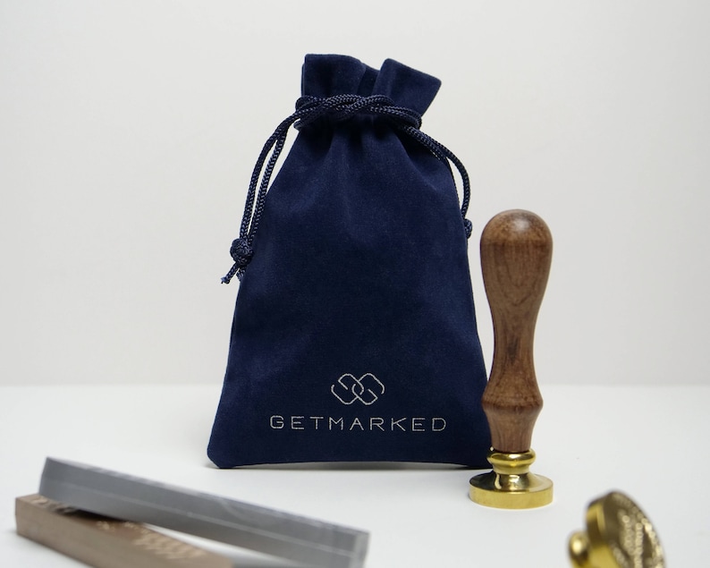 Pu&ograve; includere: Una pochette in velluto blu navy con chiusura a cordoncino e il testo "GETMARKED" in bianco. La pochette &egrave; accanto a un timbro in legno e oro con manico.