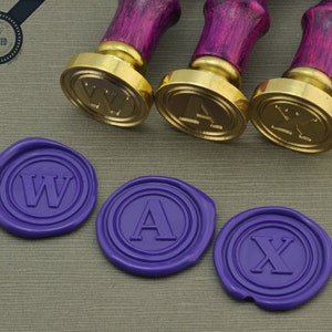 May include: Three gold-colored wax seal stamps with wooden handles. The stamps are engraved with the letters W, A, and X. Three purple wax seals with the same letters are shown below the stamps.