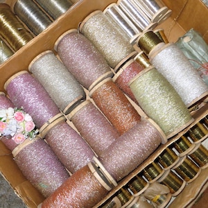May include: A close-up of a box filled with spools of metallic thread. The spools are arranged in rows and are of various colors, including gold, silver, and pink.