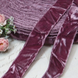 May include: A close-up shot of a roll of mauve velvet ribbon, with two lengths of ribbon in the foreground. The ribbon has a soft, textured appearance. A small floral arrangement with pink and white flowers is in the background.