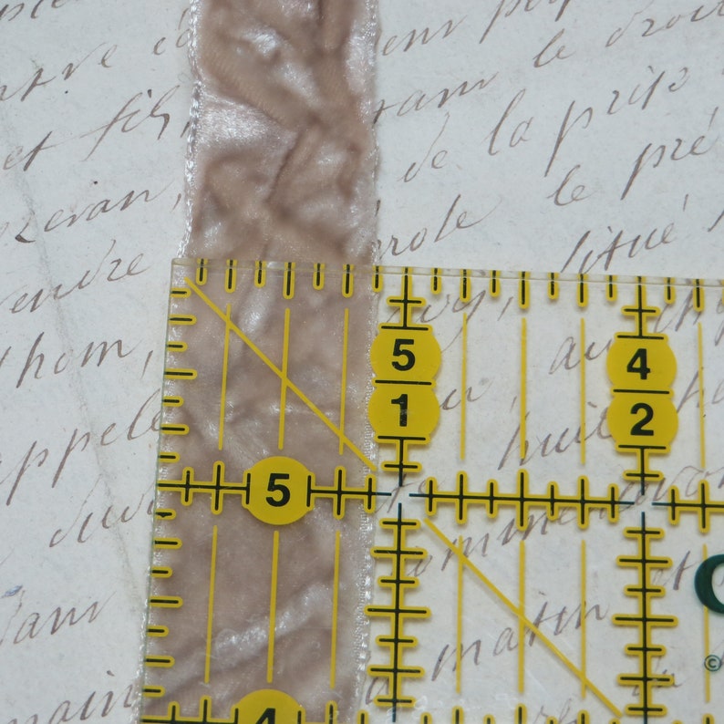 May include: A light brown velvet ribbon, approximately 2.5 cm wide, is measured with a clear ruler. The ribbon has a soft, textured appearance. The background features faded script writing.