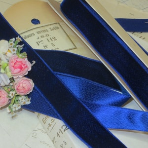 May include: Close-up of a deep blue velvet ribbon, with a soft, luxurious texture. The ribbon is partially unrolled, revealing its rich color and sheen. A vintage label and floral embellishments are also visible.