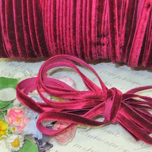 May include: A spool of deep red velvet ribbon, with a length of the ribbon unraveled and tied in a bow. The ribbon has a soft, textured appearance. The background includes floral accents and vintage paper.