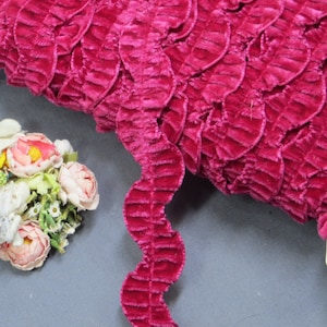 May include: A roll of vibrant fuchsia velvet ribbon with a textured, leaf-like design. The ribbon has a wavy, scalloped edge. A small bouquet of artificial flowers in shades of pink and white is in the background.