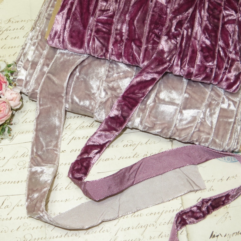 May include: A close-up shot of several rolls of velvet ribbon in shades of mauve and dusty rose. The ribbon has a soft, plush texture and a subtle sheen. The rolls are stacked, with a length of ribbon unrolled. The background is vintage paper with faded script.