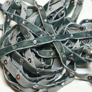 May include: A close-up of a pile of light blue velvet ribbon with silver grommets and rhinestones. The ribbon is folded and twisted, showing the details of the embellishments.