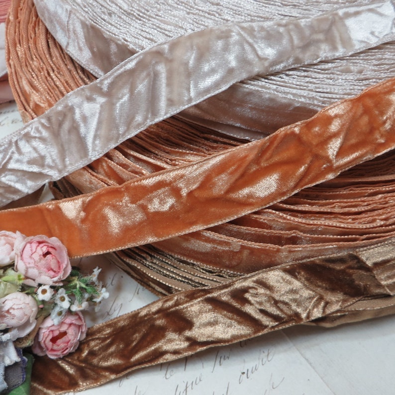 May include: A selection of velvet ribbon in shades of peach, tan, and gold. The ribbons are arranged in rolls, with the top ribbon being a light beige colour. A small bouquet of pink flowers is in the lower left corner of the image.