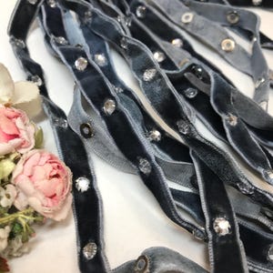 May include: A close-up of multiple strands of gray velvet ribbon with clear rhinestone embellishments. The ribbon is arranged in a loose pile, showing the texture of the fabric and the sparkle of the rhinestones.