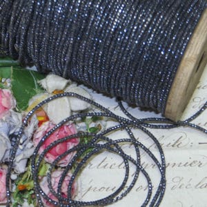 May include: A spool of dark gray, sparkly trim. The trim is wound around a wooden spool and also lies loose, creating a textured effect. The trim has a metallic sheen, reflecting light. The background includes floral elements and vintage paper.