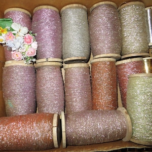 May include: A collection of spools filled with metallic thread in various colors, including pink, silver, and copper. The spools are made of wood and are arranged in a cardboard box. A small bouquet of artificial flowers is visible.