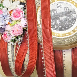 May include: Close-up of decorative ribbons and floral embellishments. The ribbons are a deep red with silver trim, and the floral arrangement features pink and white flowers. A vintage round container with a cityscape illustration is also visible.