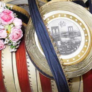 May include: Close-up of spools of red and white striped ribbon, blue ribbon, and gold trim. The spools are wrapped in white paper with a gold and black label that reads "& H& Co. Trade Mark" and features a black and white illustration of the Brooklyn Bridge.