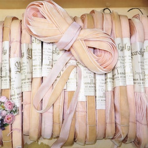 May include: A collection of vintage pink velvet ribbons, neatly rolled and packaged. The ribbons vary in shades of pink, from light blush to deeper rose tones. Some rolls have labels with text. A small bouquet of flowers is in the lower left corner.