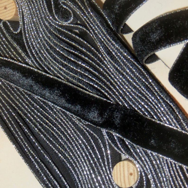 May include: Black velvet ribbon with silver metallic trim. The ribbon is folded and layered, showing the texture of the fabric.