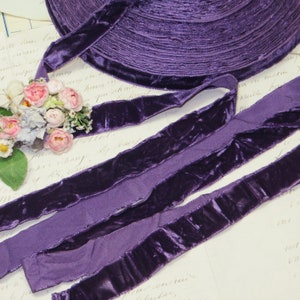 May include: A roll of deep purple velvet ribbon with a soft, lustrous texture. Several lengths of the ribbon are unrolled, displaying the rich colour and sheen. A small bouquet of pink and white flowers rests near the ribbon, adding a touch of elegance.