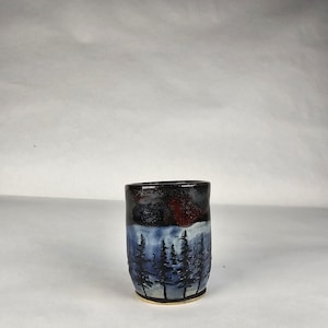 May include: A cylindrical ceramic cup featuring a hand-painted landscape design. The upper portion is a dark, glossy black, while the lower section depicts a forest of dark blue pine trees against a lighter blue background with a tan base rim.