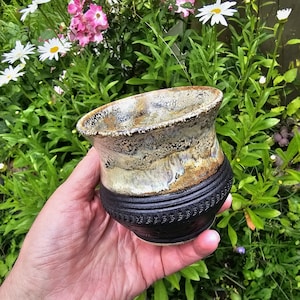 May include: A ceramic cup featuring a two-tone design with a textured matte black base and a wide, flared rim finished in a mottled cream, brown, and white glaze. The lower section of the cup displays a repeating embossed pattern around the exterior.