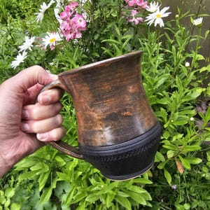 May include: A ceramic mug featuring a two-tone design with a glossy, mottled brown upper section and a matte black lower section. The base area is decorated with a textured, repeating geometric pattern that encircles the bottom of the vessel.