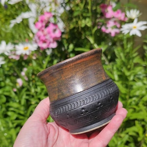 Puede incluir: Una pequeña taza de cerámica con un borde superior marrón metálico liso y un cuerpo inferior negro mate texturizado. La sección negra está decorada con patrones en relieve que incluyen filas de pequeñas hojas, puntos y líneas en zigzag.