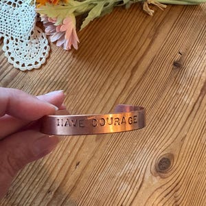 May include: A copper cuff bracelet with the words "HAVE COURAGE" engraved on the front. The bracelet is held in a hand, with a wooden table and flowers in the background.
