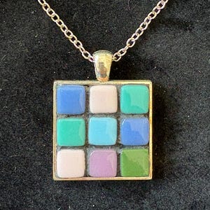 May include: A gold-toned pendant necklace featuring a square pendant with nine colorful mosaic tiles. The tiles are arranged in a 3x3 grid, with shades of blue, green, pink, and purple. The chain is a delicate link style.