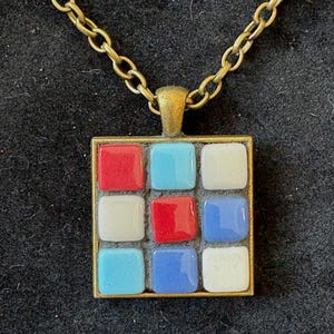 May include: A square pendant necklace with a mosaic design. The pendant features a grid of colorful squares in red, white, and blue, set in a gold-toned metal frame. The necklace has a gold-toned chain.