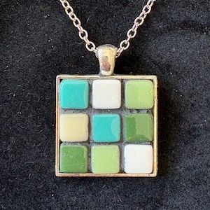 May include: A silver-toned necklace featuring a square pendant with a mosaic design. The pendant is composed of nine square tiles in shades of green, white, and teal. The chain is a delicate link style, and the pendant is set in a silver frame.
