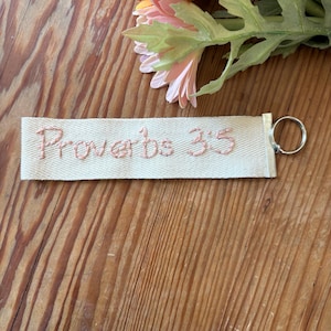 May include: A cream-colored fabric keychain with the embroidered text "Proverbs 3:5" in a light pink thread. The keychain has a silver metal ring attached. The background includes a wooden surface and colorful artificial flowers.