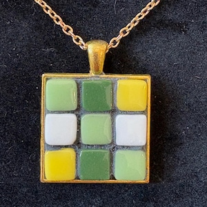 May include: A gold-toned necklace with a square pendant. The pendant features a mosaic design of nine small, square tiles in shades of green, white, and yellow. The necklace chain is also gold-toned, creating a cohesive and elegant look.