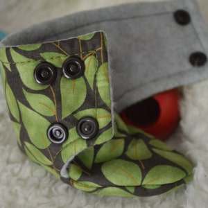 May include: Green and brown baby booties with a leaf print and four black buttons. The booties are lined with grey fleece.