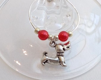 May include: A silver-colored wine charm featuring a small dog pendant, red beads, and silver accents. The charm is attached to a silver ring, designed to hang on a wine glass. The charm is on a clear wine glass.