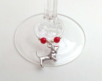 May include: A silver-colored wine charm featuring a dachshund dog pendant, red beads, and a silver ring. The charm is attached to a clear wine glass. The charm is designed to identify a wine glass.