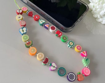 May include: A colorful phone charm with fruit-shaped beads. The charm is attached to a clear phone case with a green string.