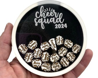 May include: A round, clear acrylic ornament with a black background and white text that reads "cheer squad 2024". Inside the ornament are small wooden cutouts in the shape of megaphones with names written on them.